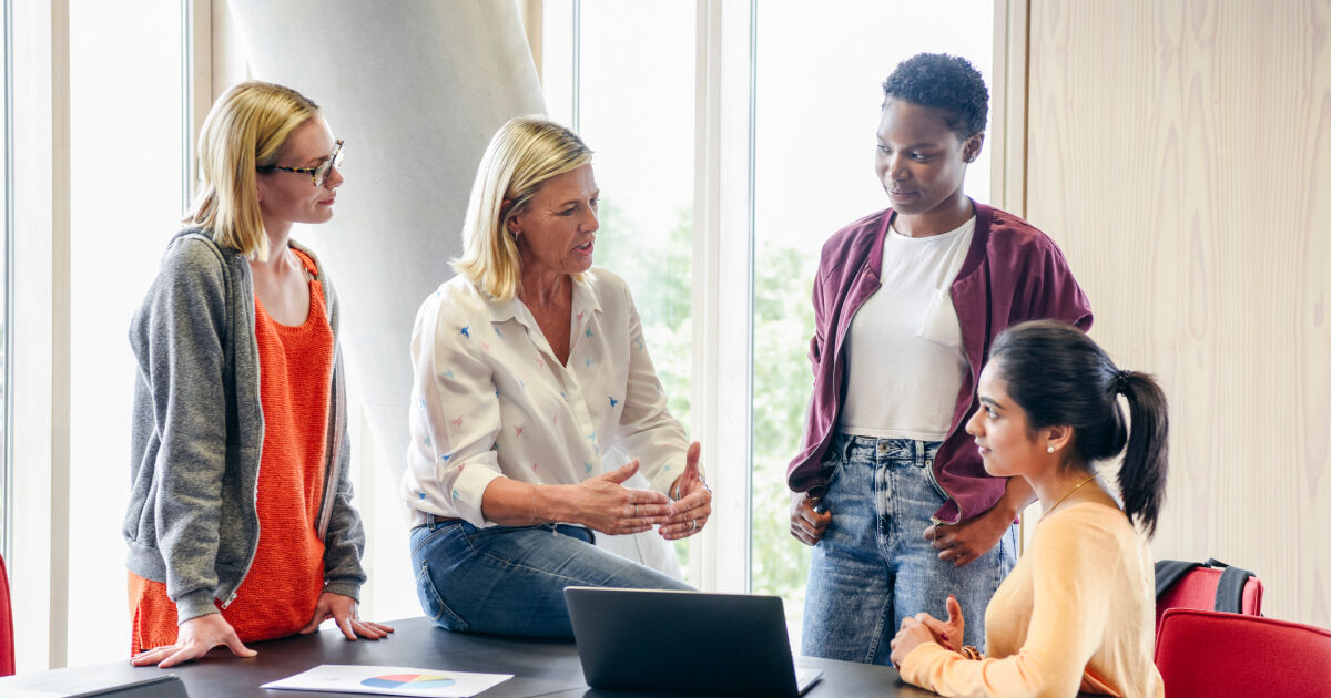 In a classroom, a teacher talks to four students. One student stands to the teacher's left; another student stands to the teacher's right; and the final student sits in a chair to the right of the second student.